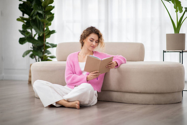 Muotokuva kaunis nuori nainen käsittelyssä kirjan rentoutuessaan kotona, rauhallinen tuhatvuotinen nainen istuu lattialla olohuoneessa ja nauttia suosikki romaani. Vapaa-aika, kirjallisuus ja ihmiset käsite - Valokuva, kuva