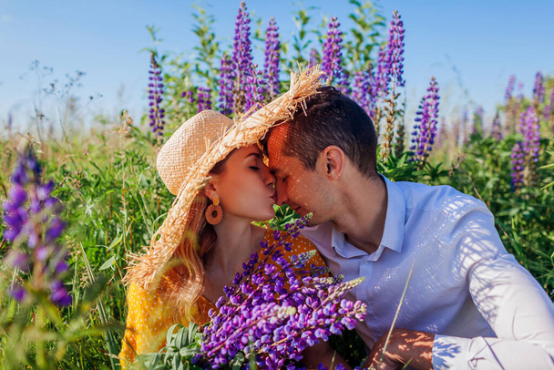Loving couple hugging in lupin field with bouquet of purple flowers. Happy woman kisses man's nose relaxing on meadow. Summer vacation - Photo, Image