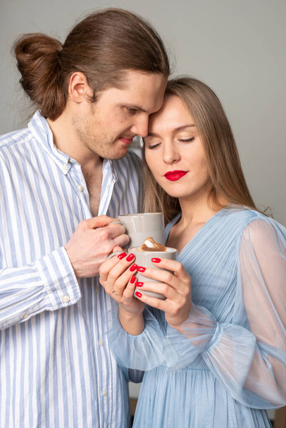 Jong lachend en gelukkig stel in lichtblauwe kleren genietend van koffie. Een gelukkige man en een mooie, stralende vrouw met rode lippen en nagels en een ring om haar vinger. Kopieerruimte - Foto, afbeelding