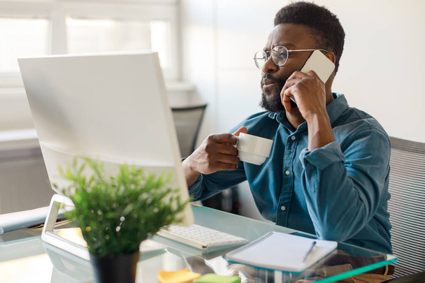 Remote business. African american male ceo talking on cellphone and using computer, male having corporate phone conversation call and drinking coffee - 写真・画像