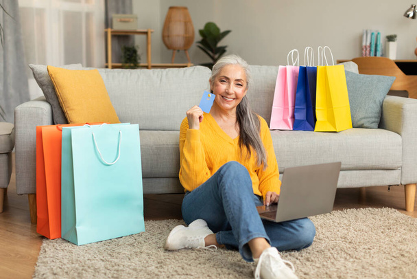 Smiling caucasian old lady with a lot of bags from store sits on floor enjoys online shopping, recommends credit card, use laptop in living room interior. Huge sale, banking and finance for order - Photo, Image