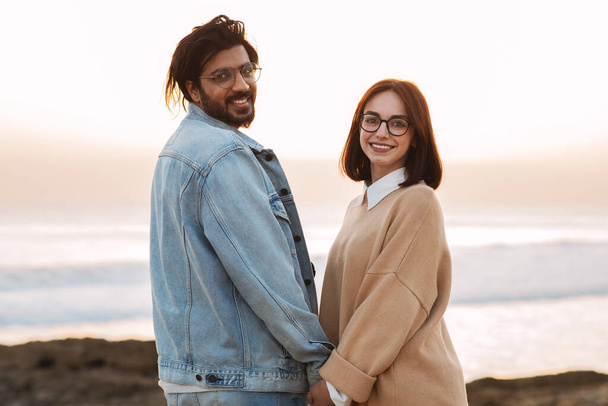 Smiling young multiethnic man and woman in casual and glasses enjoy travel, walk, date on beach near ocean, look at camera, outdoor. Trip, picnic, vacation together and active lifestyle - Fotó, kép
