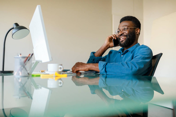 Κινητή επιχειρηματική έννοια επικοινωνίας και επιχειρηματικότητας. Μαύρος επιχειρηματίας μιλάει στο κινητό μπροστά στον υπολογιστή, κάθεται στο χώρο εργασίας στο γραφείο - Φωτογραφία, εικόνα