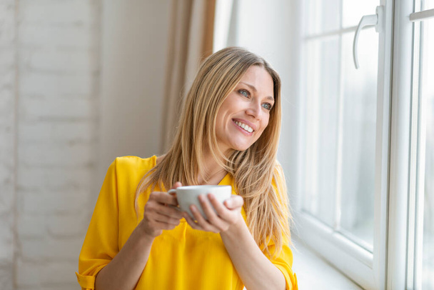 Feliz feliz muito millennial branco dona de casa loira em roupas amarelas com xícara de café gosta de beber sozinho, olha para fora da janela no interior do quarto. Emoções, descanso, relaxamento, pausa em casa, covid-19 - Foto, Imagem