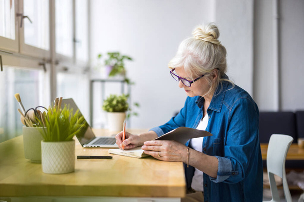 Sonriente mujer de negocios madura escribiendo en cuaderno mientras está sentada en la mesa en la oficina - Foto, Imagen