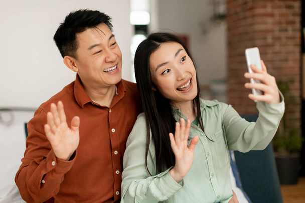 Portrait of cheerful asian married spouses using cellphone video calling and waving hello to phone webcamera, chatting with friends or family, sitting at home - Fotó, kép