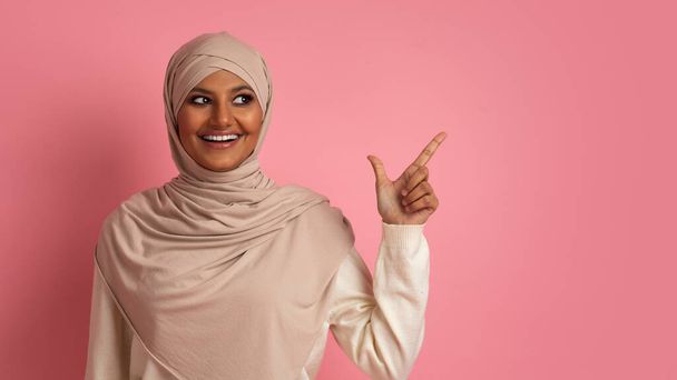 Nettes Angebot. Lächelnde Araberin im Hijab zeigt mit dem Finger auf Kopierraum, während sie über rosa Studiohintergrund steht, fröhliche Muslimin demonstriert freien Platz für Werbung - Foto, Bild
