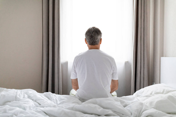 Depresión, crisis de mediana edad, crisis mental. Vista trasera del hombre de mediana edad de pelo gris con pijama sentado solo en la cama después de despertarse en casa, mirando a través de la ventana, espacio para copiar - Foto, imagen