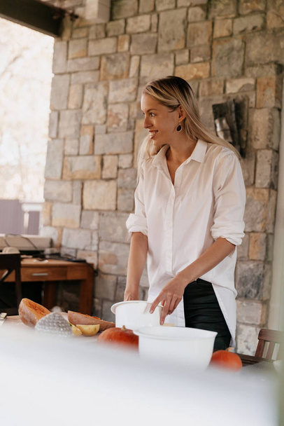 Молодая счастливая европейская женщина носит белую рубашку, держа миску и готовит еду на открытом воздухе на деревянном столе смотрит в сторону. Концепция приготовления пищи  - Фото, изображение