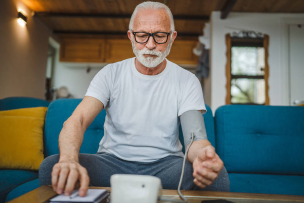 One man senior caucasian male with beard use blood pressure device to check and measure results while sit at the table at home alone real people copy space health care concept - Photo, Image