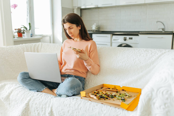 young woman is hard at work on her laptop while enjoying some pizza in her comfortable living room at home. In this real and candid moment, a young woman studies on her laptop while enjoying - Foto, immagini