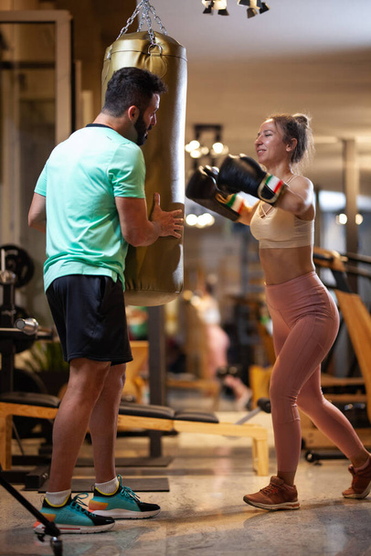 Fit girl with boxing gloves hitting a punching bag. Male fitness instructor with a female client practicing kickbox in a fitness studio. - Zdjęcie, obraz