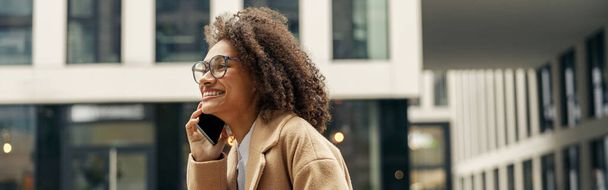 Mulher afro-americana positiva usando óculos conversando telefone com amigos durante a caminhada na cidade - Foto, Imagem