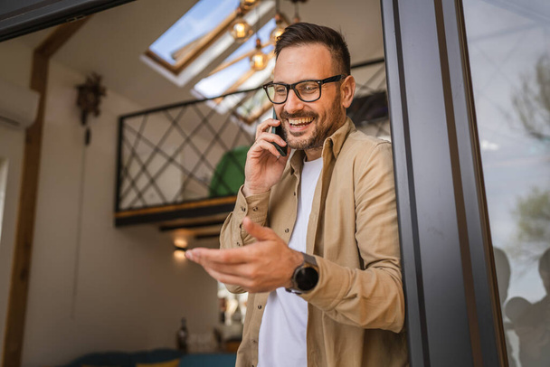 Egy férfi felnőtt kaukázusi férfi szakállal és szemüveggel áll az ajtó előtt napsütéses napon az erkélyen boldog mosoly használja a mobiltelefont, hogy egy hívást beszéljen másolás tér valós emberek - Fotó, kép