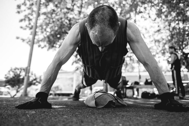 Gezond en actief: leeftijdloze en gespierde oudere man met zandgewichten training in het street workout park in de ochtend (in zwart-wit) - Foto, afbeelding