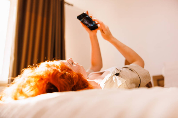 Cheerful lady with curly orange hair is sitting on the bed talking on video call with her boyfriend. High quality photo - Foto, Bild