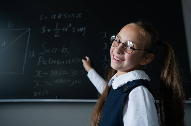 Portrait of a caucasian girl in glasses in the classroom. The schoolgirl writes the formula with chalk on the blackboard and looks into the camera - Φωτογραφία, εικόνα