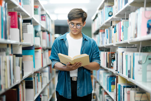 Νεαρός φοιτητής με casual ντύσιμο με σακίδιο πλάτης που στέκεται, διαβάζει ή επιλέγει βιβλία για να διαβάσει στη βιβλιοθήκη για σπουδές, ακαδημαϊκή έρευνα ή σχολικές εργασίες. Μάθηση, Εκπαίδευση έννοια. - Φωτογραφία, εικόνα