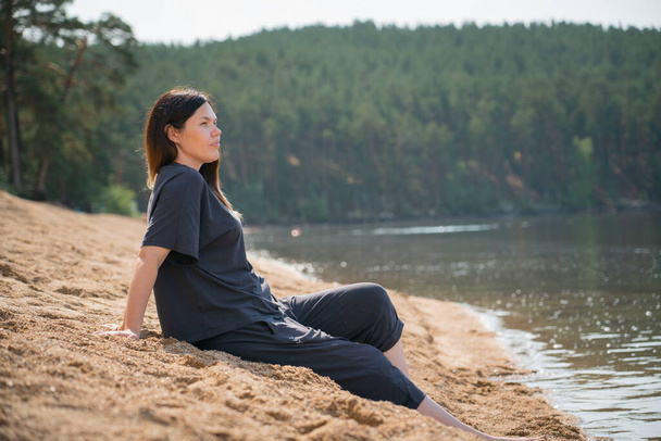 A young pregnant woman sits on the beach near the water and admires the view. Unity with nature, alone with yourself, grounding, harmony, stress relief, relaxation, mental healing. High quality photo - Photo, Image