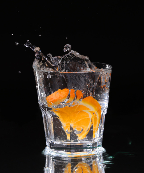 Slice of lemon splashing into a glass of water with a spray of water droplets in motion suspended in the air above the glass on a dark background. - Foto, imagen