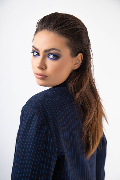 Beauty. Portrait of face of beautiful brunette with dark long hair combed back, bright blue makeup, looking through shoulder into camera. Close-up. Studio, white background. Emancipation. Fashion.  - Photo, image