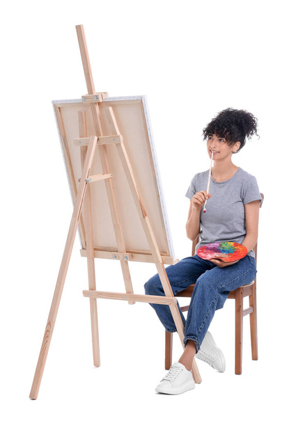 Young woman holding brush and artist`s palette near easel with canvas against white background - Fotó, kép
