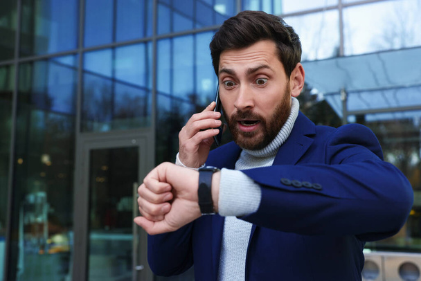 Hombre de negocios emocional hablando en el teléfono inteligente y mirando el reloj cerca del edificio. Llegar tarde concepto - Foto, Imagen