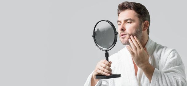 Millennial hispanic man looking in mirror in bathroom, facial skin and stubble. Male beauty care product. Skincare, home spa. Beauty portrait of a beautiful man. Spa, moisturizing nourishing creme - Photo, Image