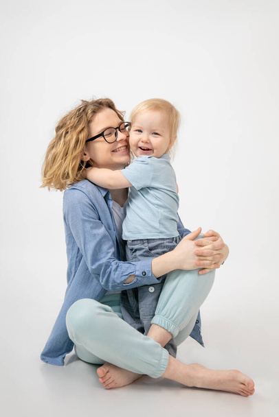 Happy young red-haired woman is sitting on white background in studio, laughing and hugging her little daughter. They are dressed in clothes of same shades. Motherhood. Parents and children.  - Фото, зображення