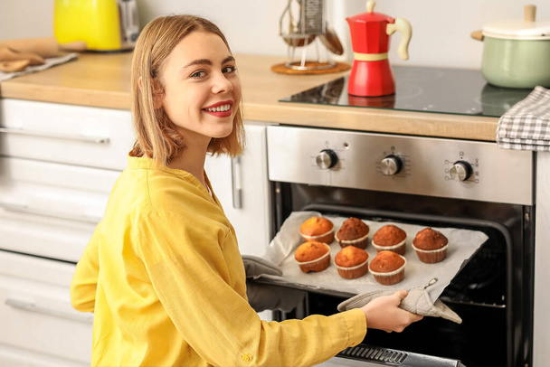 Young woman taking tray with cupcakes from oven in kitchen - 写真・画像