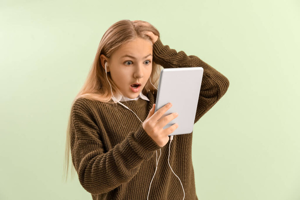 Shocked little girl in earphones using tablet computer on green background - Foto, afbeelding