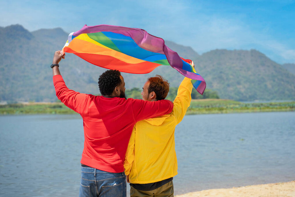 Back view. African gay and asian gay couple love moment spending good time together, raising and waving the LGBT flag on the beach - Фото, зображення