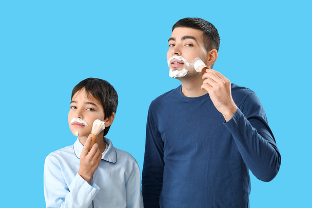 Человек и его маленький сын наносят пену для бритья на лица на синем фоне - Фото, изображение