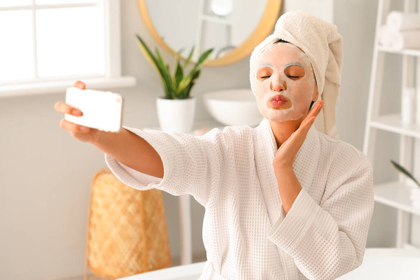 Mujer joven con máscara de sábana tomando selfie en el baño - Foto, Imagen