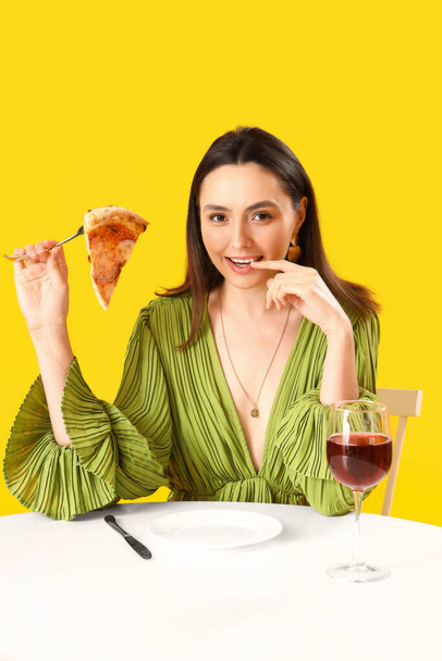 Sarı arka plandaki masada lezzetli pizzası ve bir bardak şarabı olan genç bir kadın. - Fotoğraf, Görsel