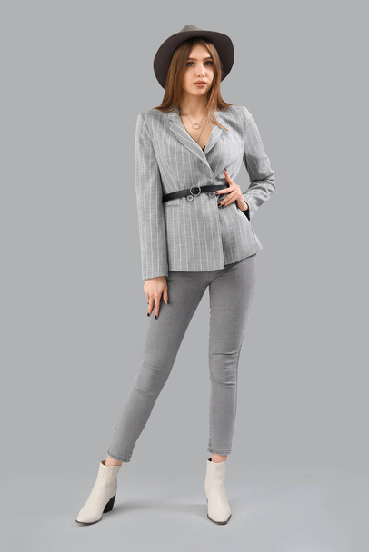 Stylish young woman in jacket, jeans and hat on grey background - Фото, изображение
