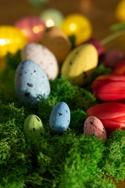 Wielkanocne kolorowe jajka na zielonej trawie wokół tulipanów i drewnianej deski Happy Easter. Wysokiej jakości zdjęcie - Zdjęcie, obraz