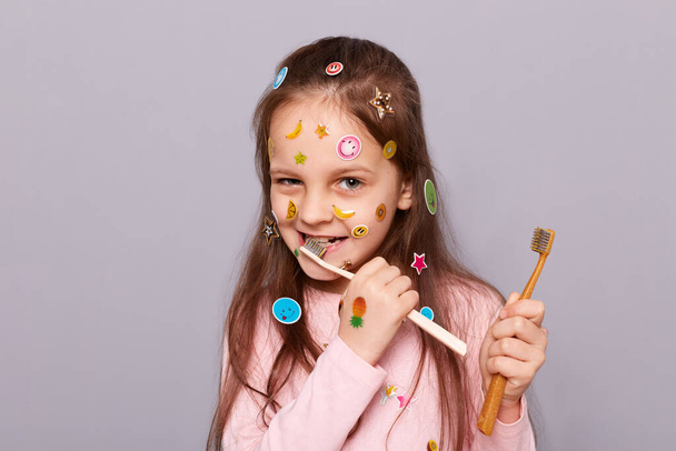 Portrait of funny playful little girl covered with colorful stickers wearing pink shirt standing isolated over gray background, brushing her teeth, taking care of her mouth alone, dental problems. - Fotoğraf, Görsel