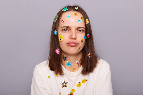 Indoor shot of sad stressed offended disappointed woman covered with funny stickers looking at camera with pout lips, being upset, posing isolated over gray background - Foto, Bild