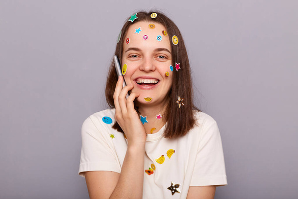 Photo of positive joyful beautiful woman covered with funny stickers holding smartphone and talking with friends, laughing joyfully, posing isolated over gray background - 写真・画像