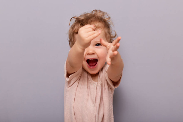 Portrait of playful cheerful cute little toddler girl standing isolated over gray background, looking at camera and outstretched hands, yelling with happiness. - Φωτογραφία, εικόνα