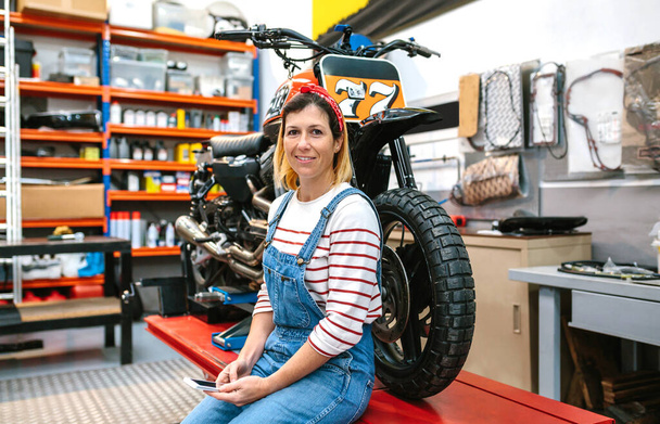 Portrait of smiling mechanic woman looking at camera while holding phone in her hands sitting over platform with custom motorcycle on factory - Fotografie, Obrázek