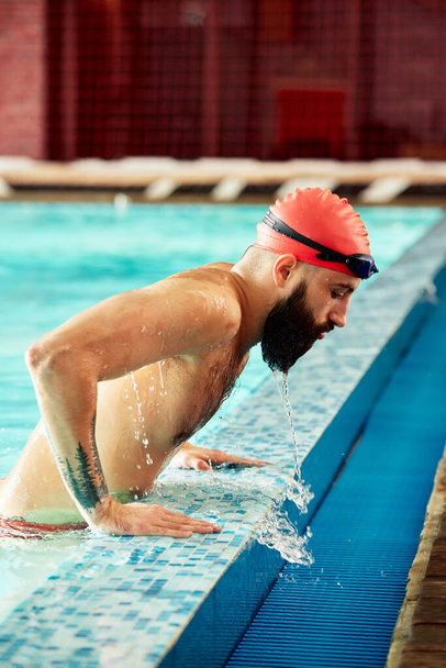 Un nadador en un sombrero y gafas en el lado de la piscina, un nadador masculino emerge de la piscina en una gorra roja para nadar, volando fuera de la piscina emerge de las vacaciones de entrenamiento de natación. - Foto, Imagen