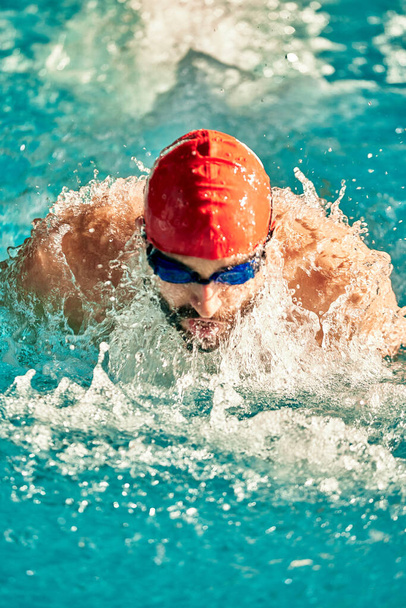 Zwemmen - mannelijke zwemmer zwemmende schoolslag. close-up portret van de man doet borst beroerte zwemmen in zwembad het dragen van rode badmuts en badbril. - Foto, afbeelding