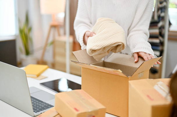 Imagen de primer plano de una vendedora de ropa de moda femenina empacando un producto de envío en una caja de cartón, trabajando en su oficina en casa. - Foto, Imagen
