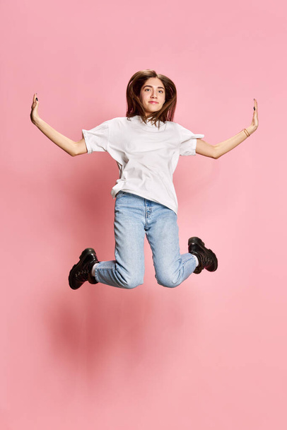 Full-length portrait of young, beautiful, brunette girl in casual clothes jumping against pink studio background. Relaxation. Concept of youth, emotions, facial expression, lifestyle - Foto, Bild