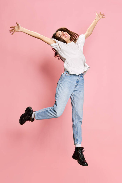 Portrait of smiling, young girl in casual jeans and t-shirt posing in a jump, spreading hands in happiness against pink background. Youth, emotions, facial expression, lifestyle, celebration concept - Photo, Image