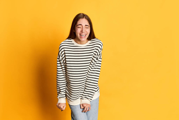 Portrait of young beautiful girl in striped sweater smiling, laughing against yellow studio background. Funny jokes and good mood. Concept of youth, emotions, facial expression, lifestyle - Foto, immagini