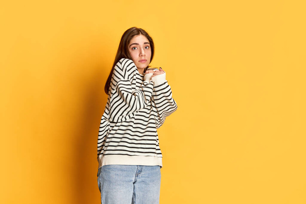 Studio portrait of young girl in stripes sweater posing with little fear in eyes agaisnt yellow backgorund. Feeling shy and uncertain. Concept of youth, emotions, facial expression, lifestyle - Fotó, kép