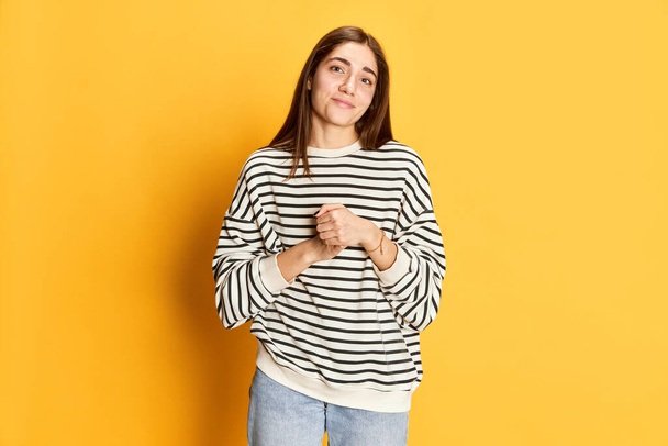 Portrait of cute looking, attractive youn g girl calmly posing in striped sweater against yellow studio background. Concept of youth, emotions, facial expression, lifestyle - 写真・画像